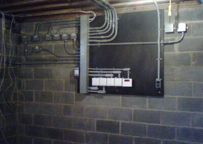 Electrical panel with multiple cables mounted on a concrete wall in a basement.