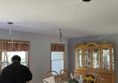 A dining room with a wooden table, chairs, and a china cabinet. A person works on electrical wiring hanging from the ceiling. Tools and materials are on the table.