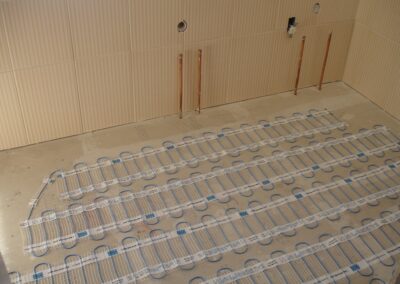 Electric underfloor heating system installed on a concrete subfloor with rows of heating mats and visible copper pipes in an unfinished room.