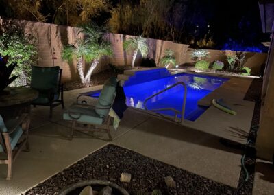 A dimly lit backyard with a pool surrounded by chairs and plants. The pool is illuminated with blue lights.