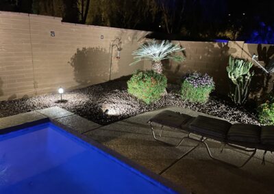 Nighttime backyard scene with illuminated pool, patio chaise lounge, and garden plants against a brick wall.
