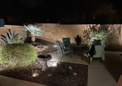 Nighttime patio scene with chairs and a table. Surrounded by desert plants, a lit up birdbath, and a brick wall.
