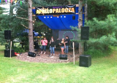 Outdoor music stage with a group of people singing and playing instruments, surrounded by trees. A sign above reads "Apollopalooza." Speakers and blue tarp above complete the setup.