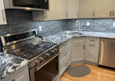 Modern kitchen with stainless steel appliances, gray cabinets, marble countertops, and a stainless steel sink. A kettle is on the gas stove. Gray rug on the wooden floor.