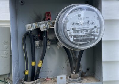 An open electrical meter box mounted on a white exterior wall, displaying wires and a round meter with labels and numbers.
