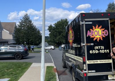 Black utility truck labeled "Apollo Electric" parked on a suburban street near a lamppost, with cars and houses visible in the background.