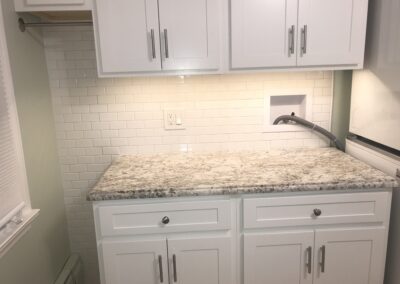 A kitchen features white cabinets with metal handles, a granite countertop, and a white subway tile backsplash. A small recessed space contains an outlet and a coiled cable.