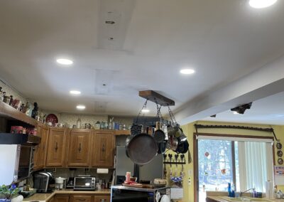 A kitchen with wooden cabinets, hanging pots, a microwave on the counter, and recessed ceiling lights. The ceiling shows a partially repaired section.