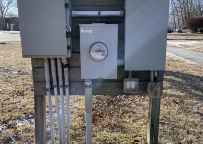Outdoor electrical meter and panels mounted on a wooden board with conduit pipes leading into the ground, located in a grassy area with some snow.