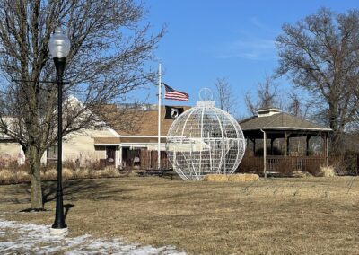 A snowy yard features a spherical wire structure, gazebo, and building. A streetlamp and trees are present, with an American flag flying in the background under a clear blue sky.
