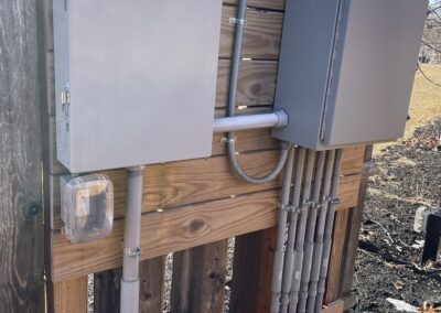 Two gray electrical panels mounted on a wooden fence with connected conduits in an outdoor setting. Snow and dry leaves on the ground.