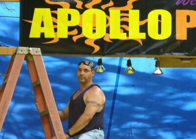 A man in a tank top and bandana stands on a ladder in front of a sign that reads "APOLLO" with lights beneath.