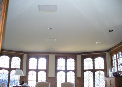 Room with large windows, wood paneling, and light fixtures on the ceiling. A table with a lamp and chairs is visible.