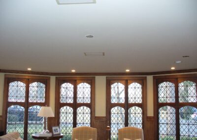 A room with a curved wall of diamond-patterned windows, two beige armchairs, a side table with a lamp, and recessed ceiling lights.