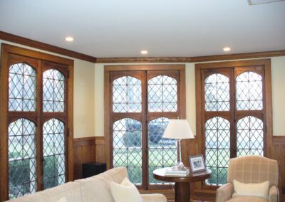 Living room with diamond-patterned leaded glass windows, beige sofa, round wooden table, and armchair. Table has a lamp and framed photo.