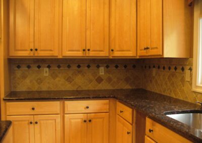 A kitchen with light wood cabinets, a tiled backsplash, and dark marble countertops. A stainless steel sink is visible on the right.