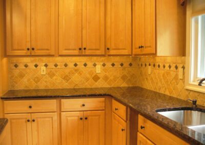 A kitchen with wooden cabinets, dark granite countertops, a tiled backsplash, and a stainless steel sink under a window.