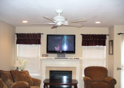 Living room with a sofa and armchair, TV mounted above a fireplace, brown curtains, and a ceiling fan.