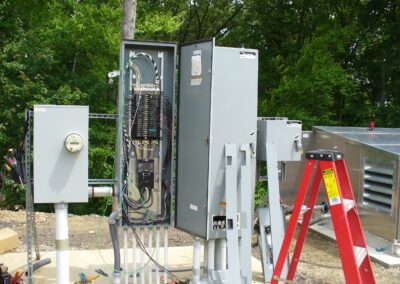 Electrical boxes with open panels and wiring outdoors. Nearby, a red ladder and ventilation unit are visible, with a background of green trees.