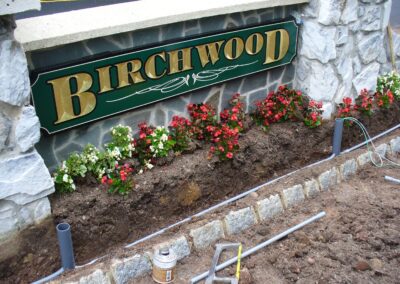 A sign with "Birchwood" on it, surrounded by stonework and red and white flowers, with exposed soil and tools visible in the foreground.