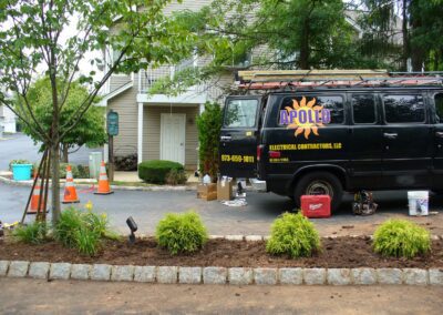 A black electrician van parked on the street near a residential building. Equipment and tools are on the ground next to it. Traffic cones are placed around the area.